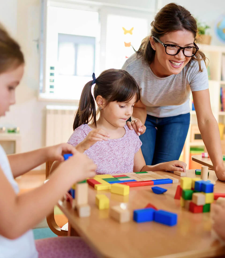 A teacher smiles while assisting children playing with colorful building blocks at a table in a bright classroom, conveying a cheerful, educational atmosphere.