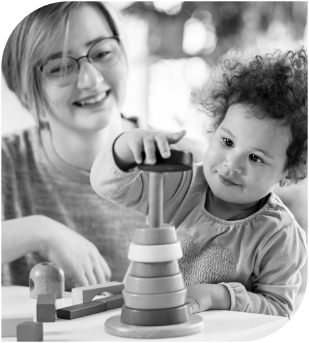 A joyful toddler stacks rings on a peg, assisted by a smiling adult. The scene conveys learning and play in a nurturing environment.