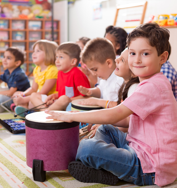 A group of children sits in a classroom, engaging with drums, focused on making music together on a colorful rug.